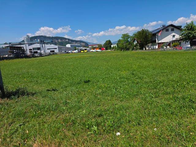 Weitläufige grüne Wiese mit Häusern und Industrieanlagen im Hintergrund, umgeben von einer Berglandschaft.