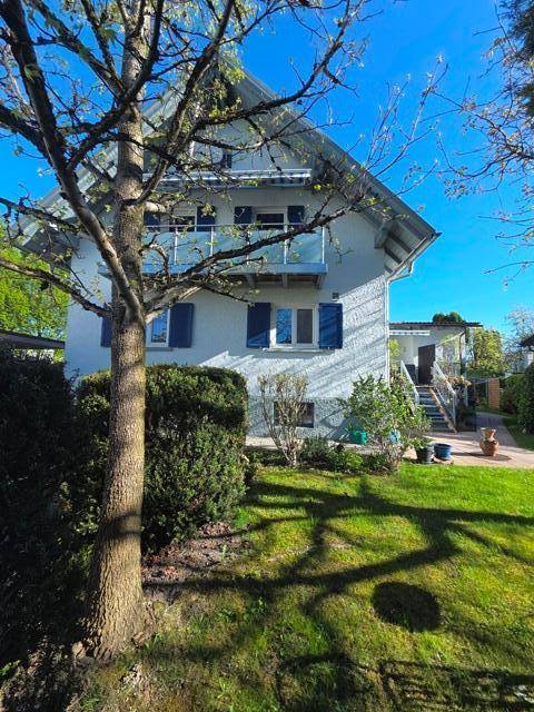 Wohnhaus mit Satteldach, Balkon und blauen Fensterläden, umgeben von einem grünen Garten.