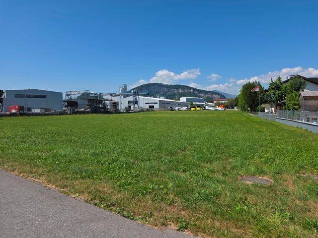 Großes grünes Feld mit Industriegebäuden und Bergen im Hintergrund unter einem klaren blauen Himmel.