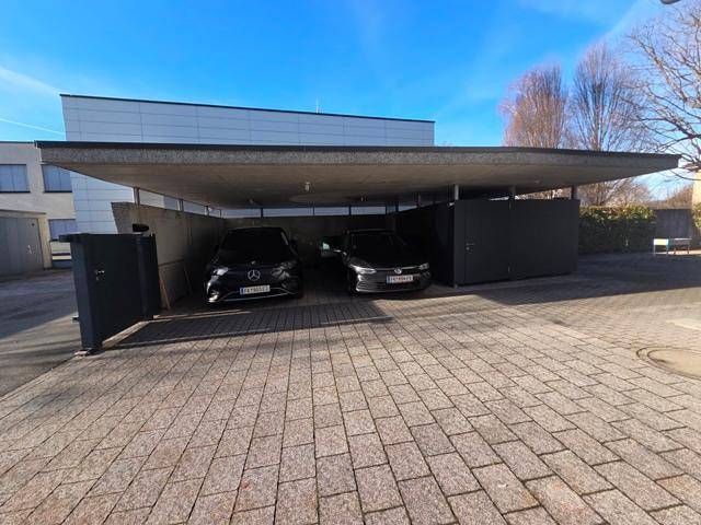 Überdachter Carport mit zwei geparkten Fahrzeugen und gepflastertem Vorplatz.