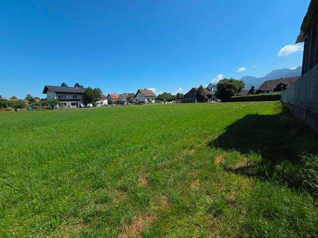 Weite grüne Fläche mit Wohnhäusern und einer Bergkette am Horizont unter strahlend blauem Himmel.