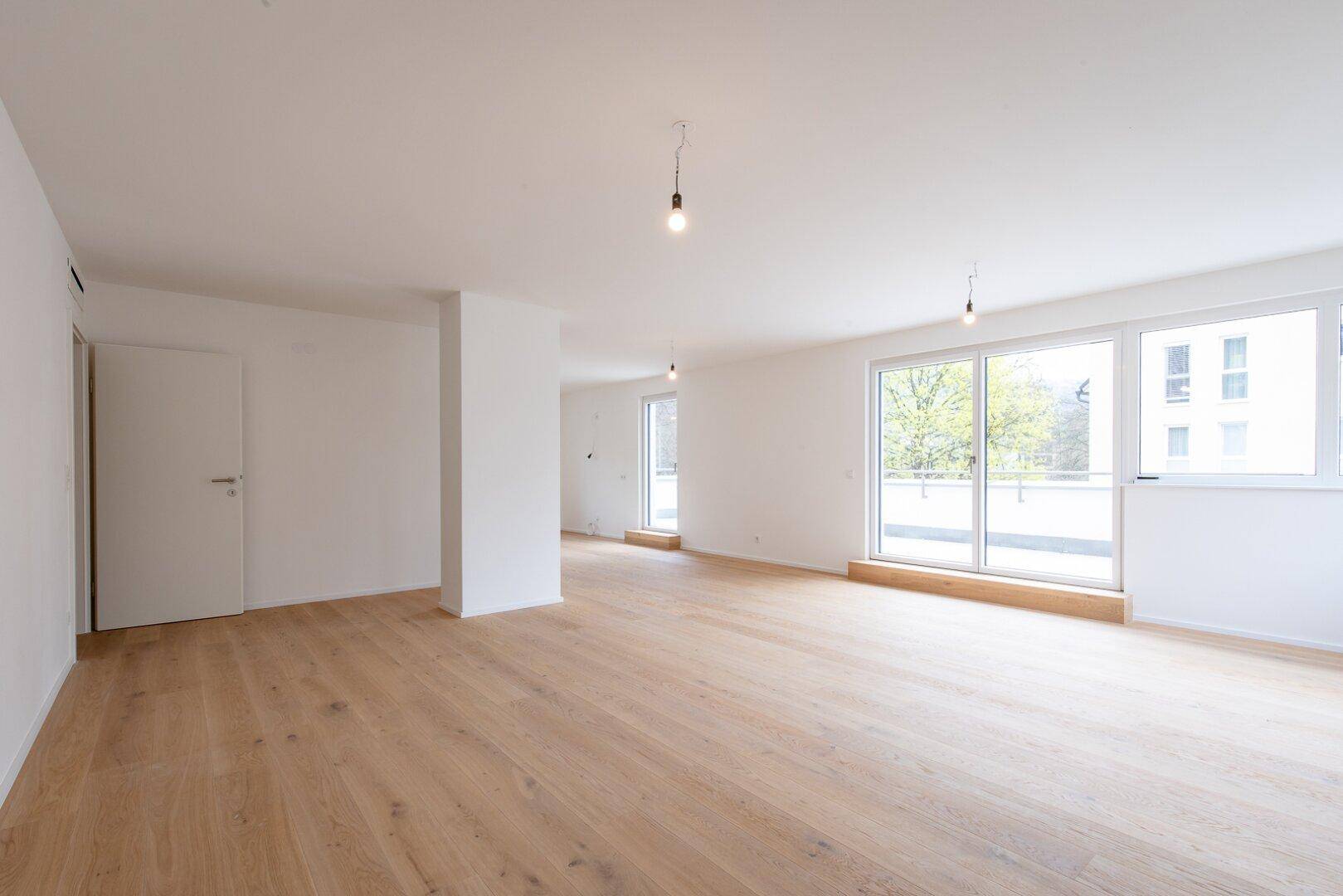 Heller, leerer Wohnbereich mit großen Fenstern, Holzboden und hohen Decken in einem Neubau.