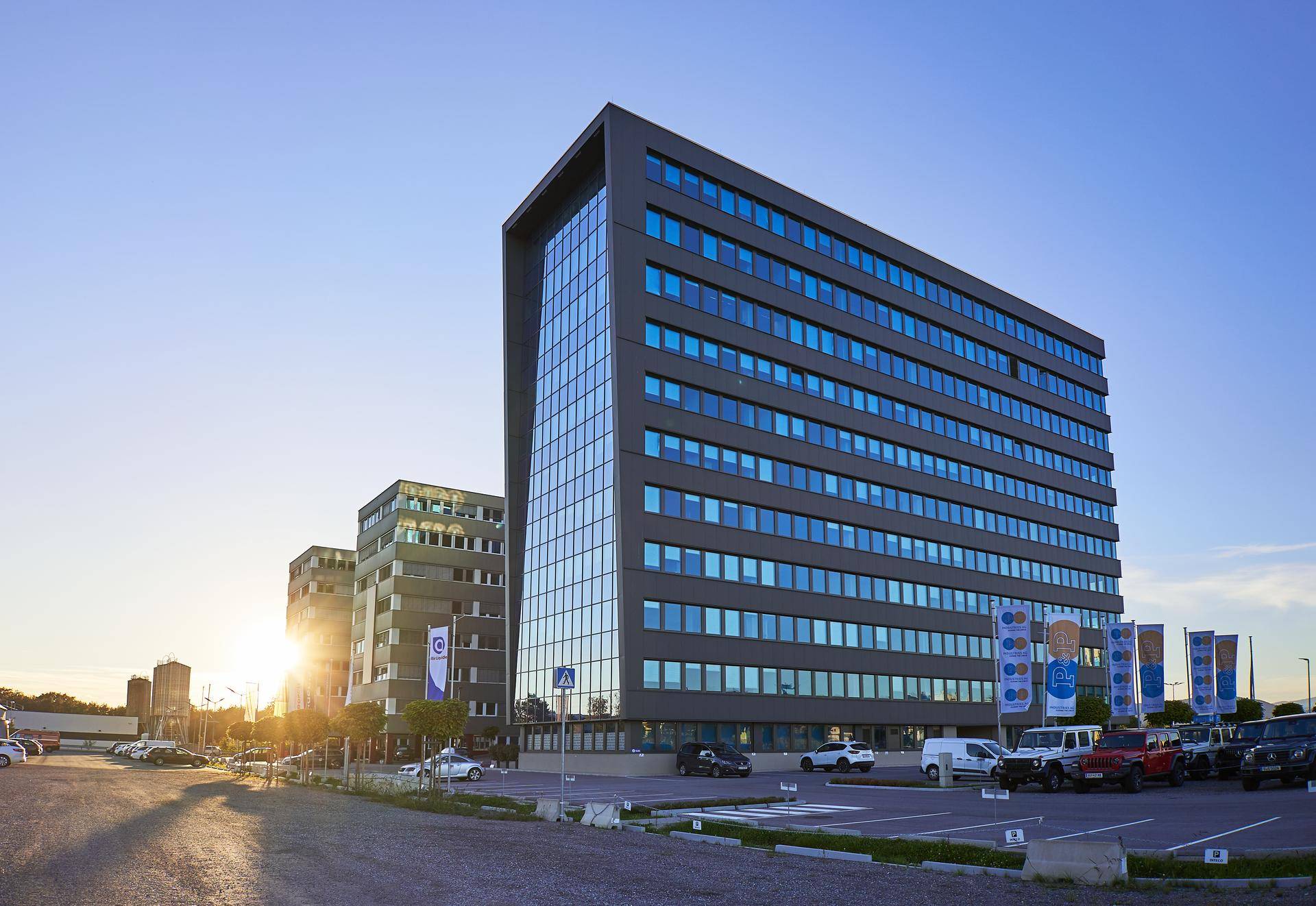 Modernes Bürogebäude mit Glasfassade und Parkplatz bei Sonnenuntergang, umgeben von weiteren Geschäftsgebäuden.