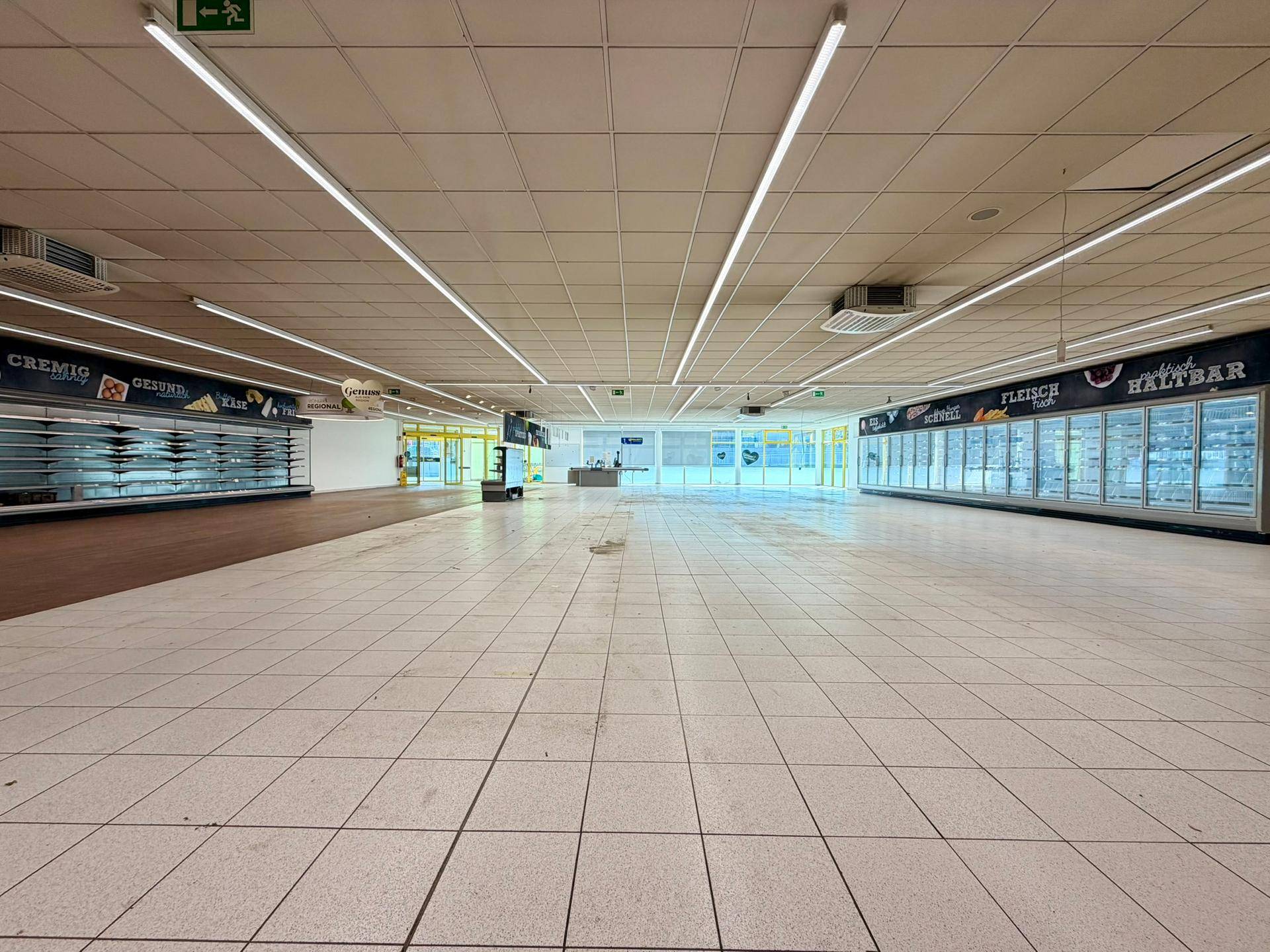 Weitläufiger, leerer Supermarkt-Verkaufsraum mit Fliesenboden und leeren Regalen.