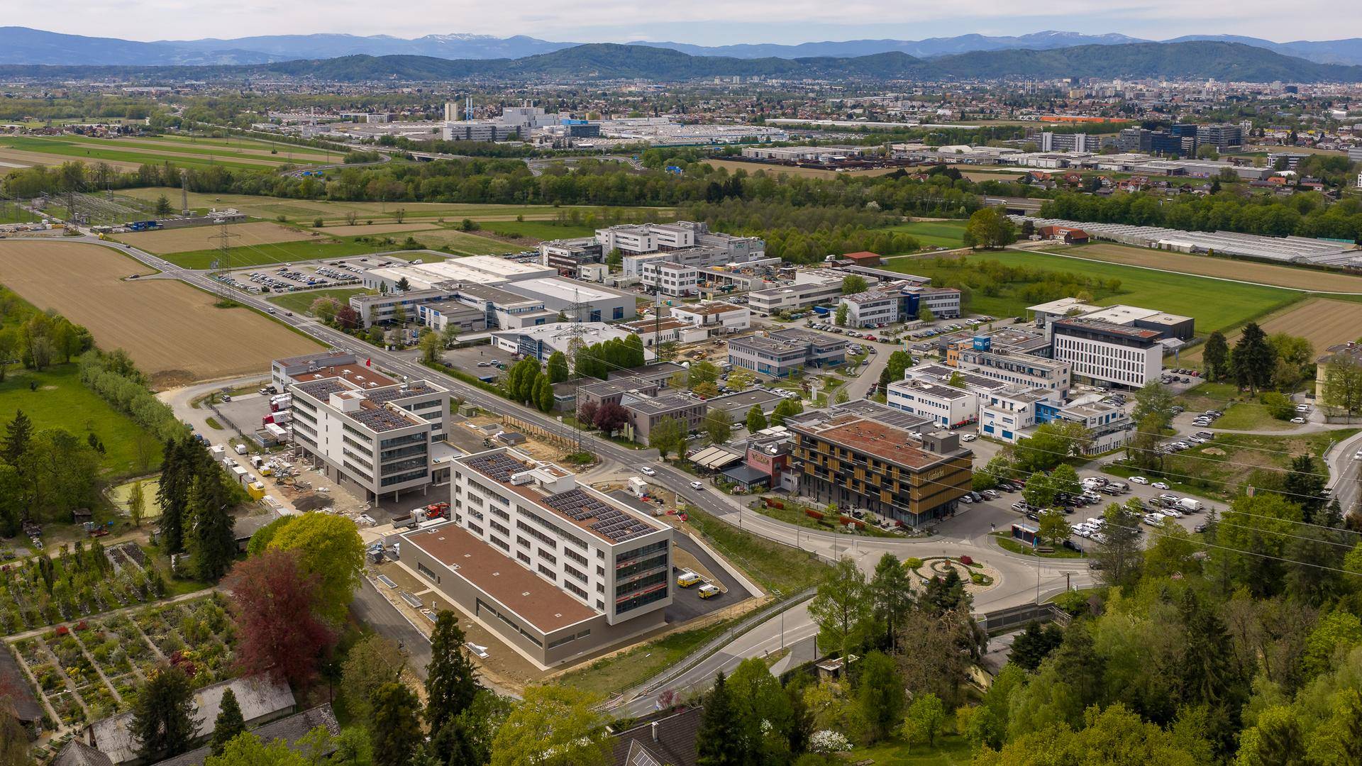 Luftaufnahme eines Gewerbegebiets mit modernen Gebäuden, umgeben von Grünflächen und Bergen.