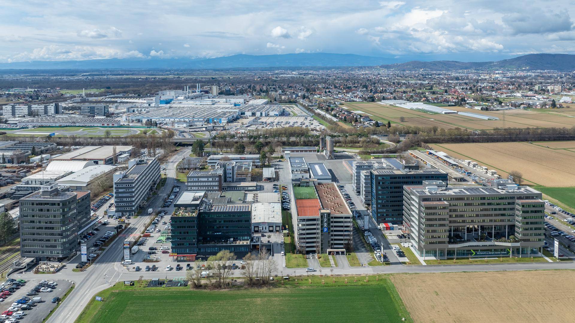 Weitläufige Luftaufnahme eines Gewerbegebiets mit mehreren Bürogebäuden, Feldern und Bergen im Hintergrund.