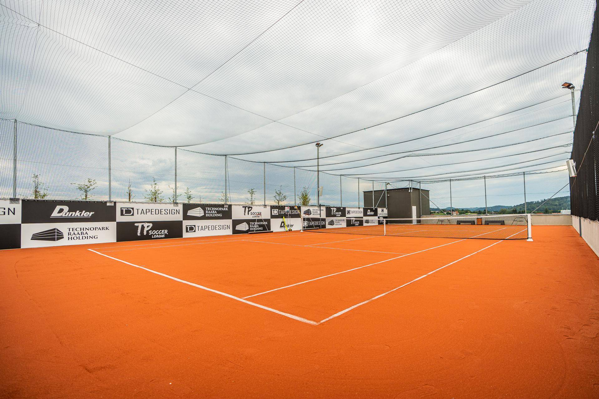 Roter Tennisplatz im Freien mit Netz und Umzäunung, bereit für ein Match unter bewölktem Himmel.