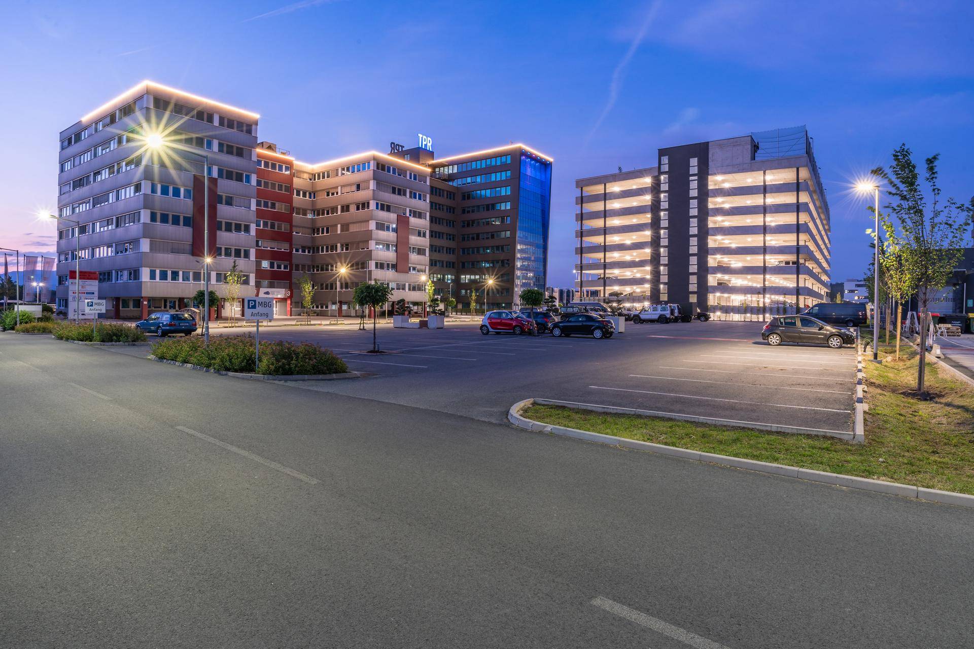 Bürogebäude bei Dämmerung mit beleuchteten Fassaden und einem großen Parkplatz im Vordergrund.