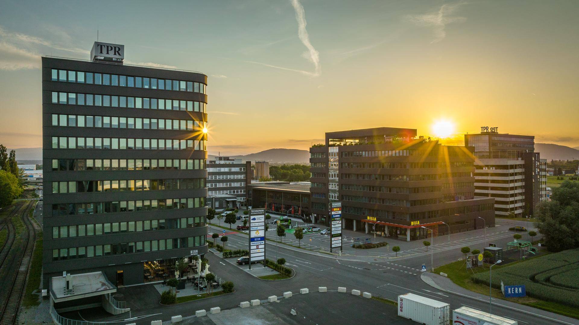 Bürogebäude in einer Gewerbezone bei Sonnenuntergang, mit Bergen im Hintergrund und einer Straße im Vordergrund.
