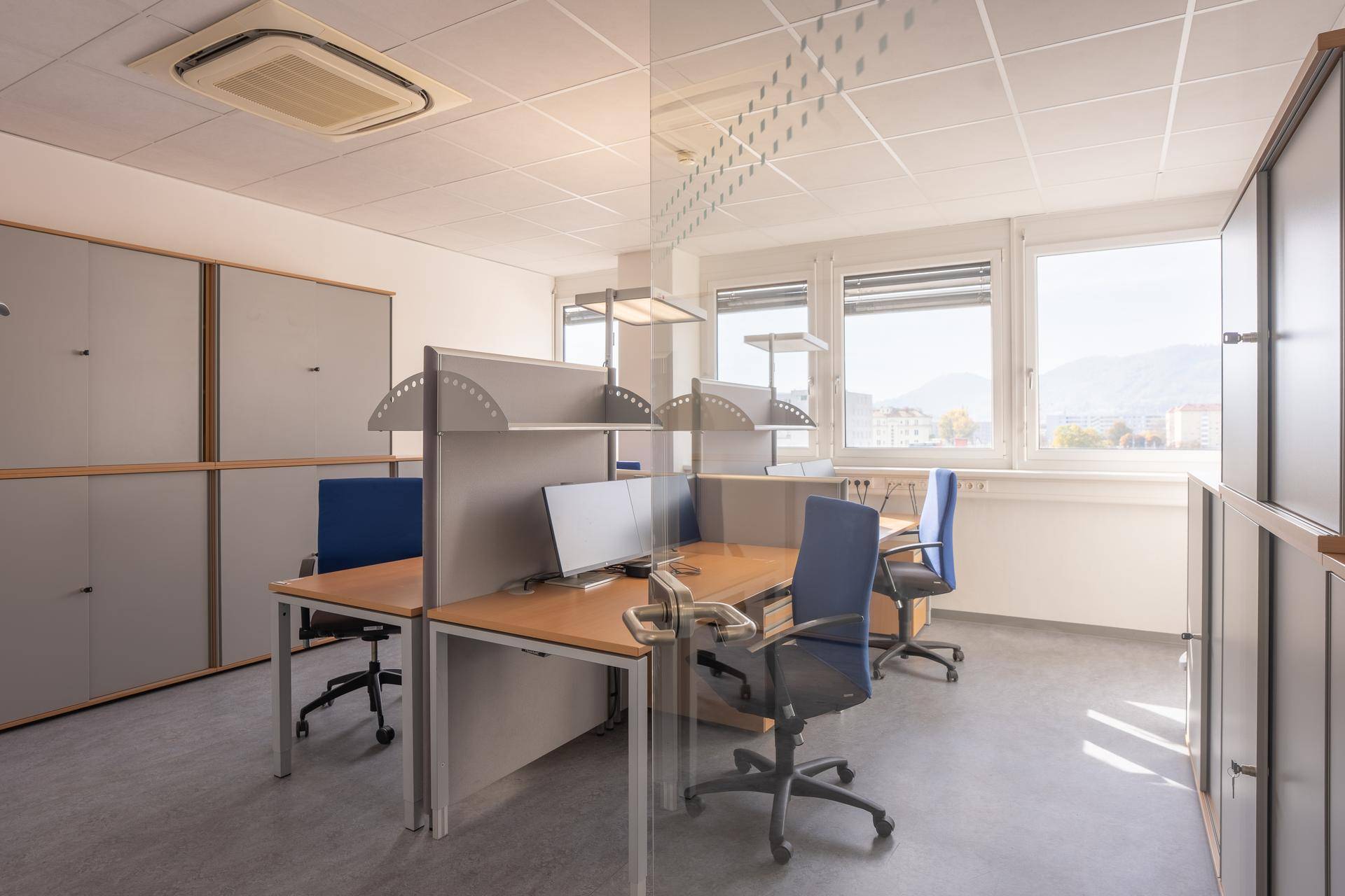 Offener Bürobereich mit mehreren Arbeitsplätzen, Stauraum und großen Fenstern mit Blick ins Grüne.