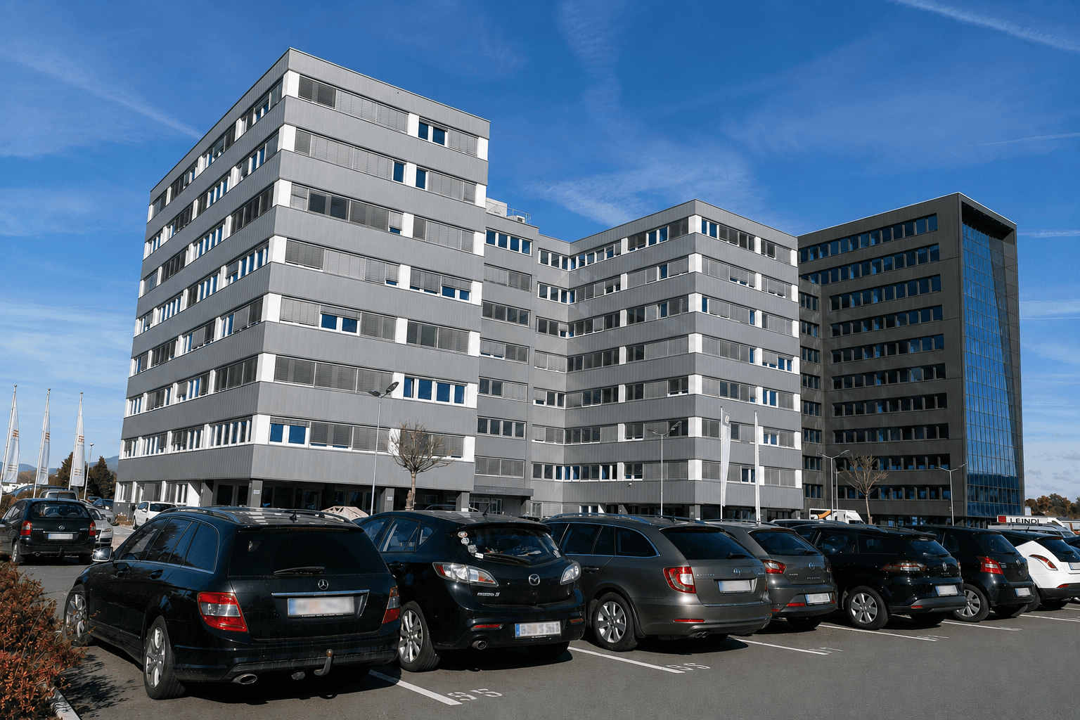 Ansicht eines modernen Bürogebäudes mit vielen Fenstern und einem großen Parkplatz unter blauem Himmel.