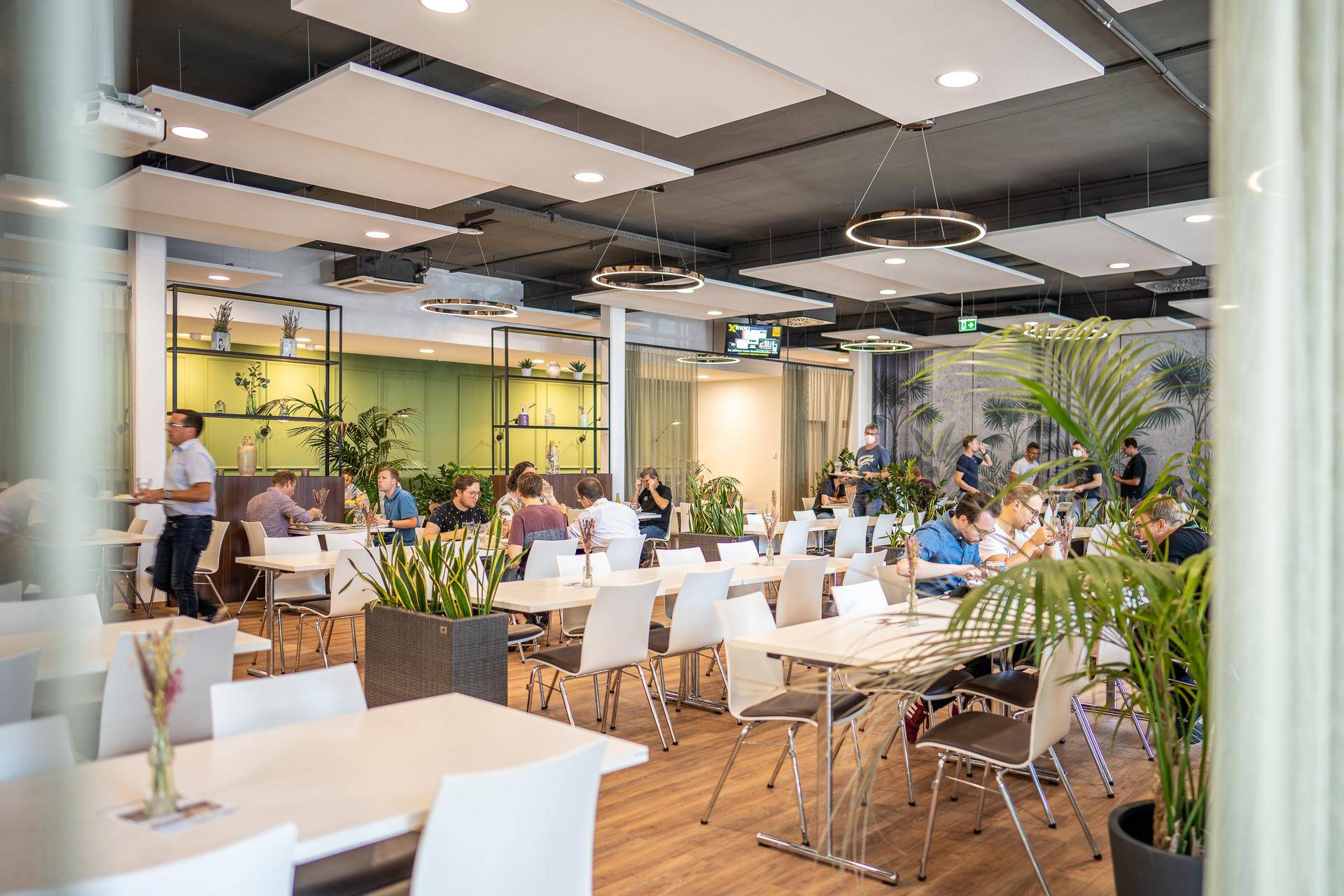 Helle und moderne Cafeteria mit vielen Tischen, Stühlen und Pflanzen, belebt mit Gästen.