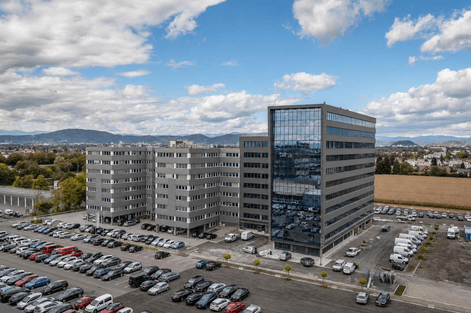 Luftaufnahme eines großen Bürokomplexes mit weitläufigen Parkplätzen und umliegender Landschaft unter bewölktem Himmel.