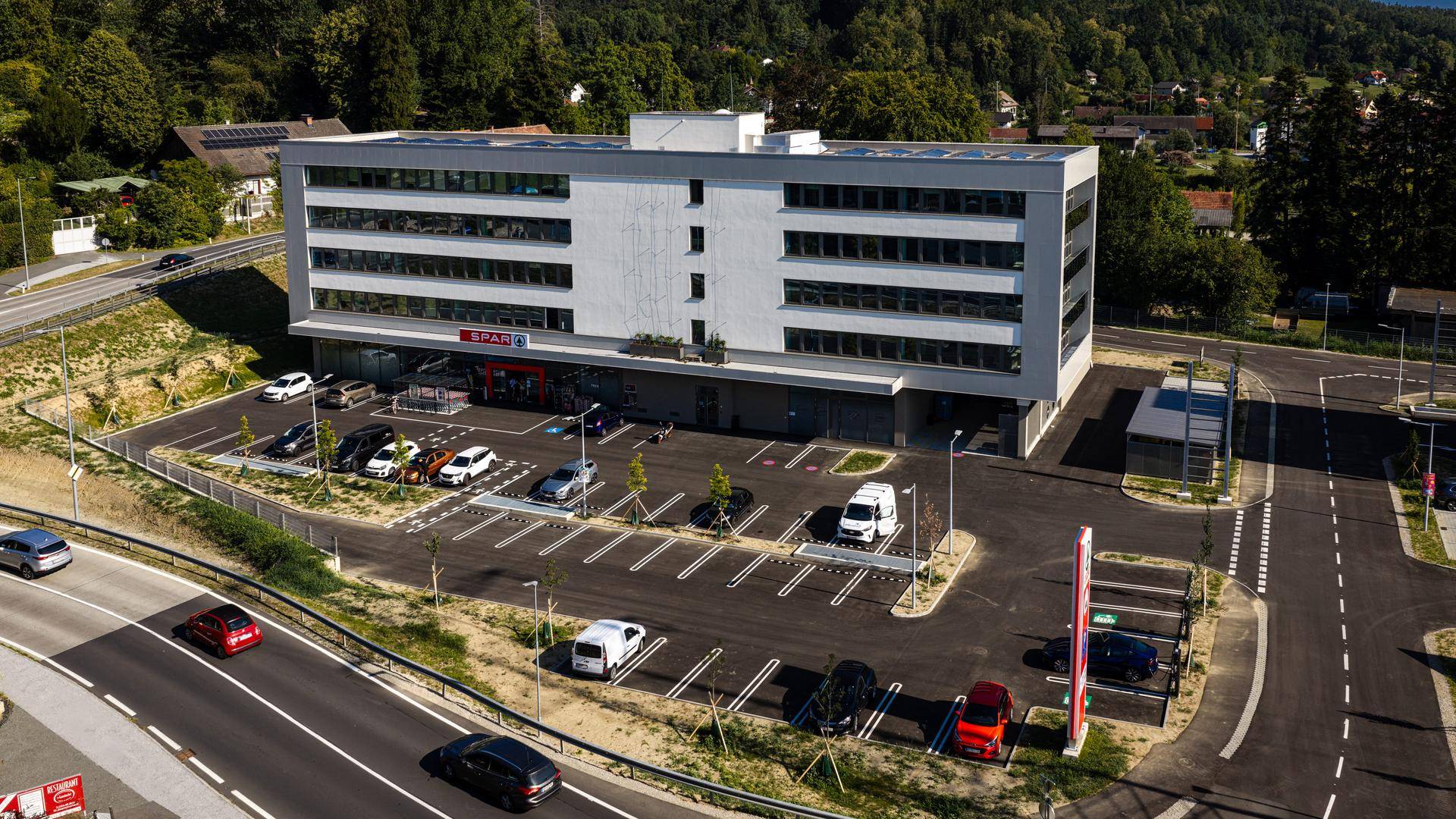 Modernes Geschäftsgebäude mit Supermarkt und großem Parkplatz in grüner Umgebung.