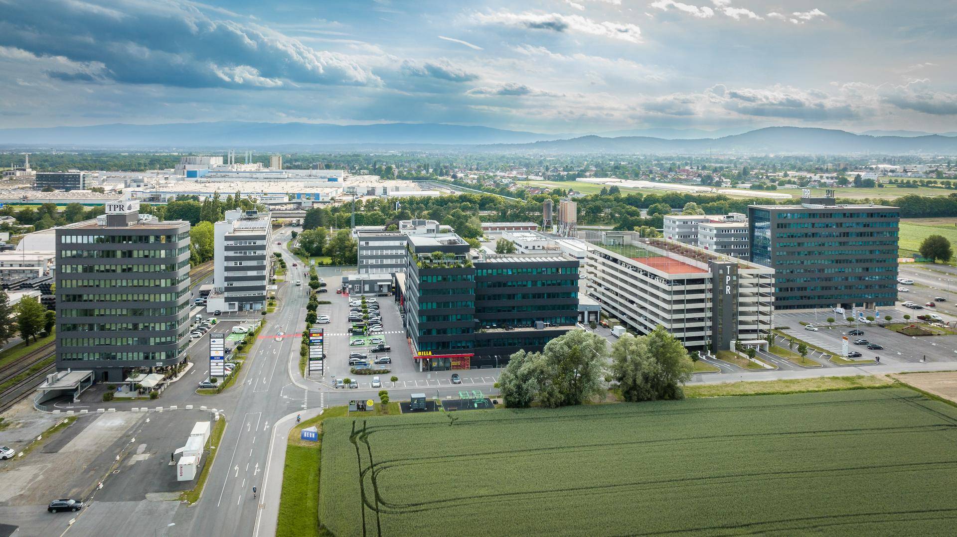Luftaufnahme eines modernen Bürokomplexes mit mehreren Gebäuden, Parkplätzen und angrenzenden Feldern, im Hintergrund Berge.