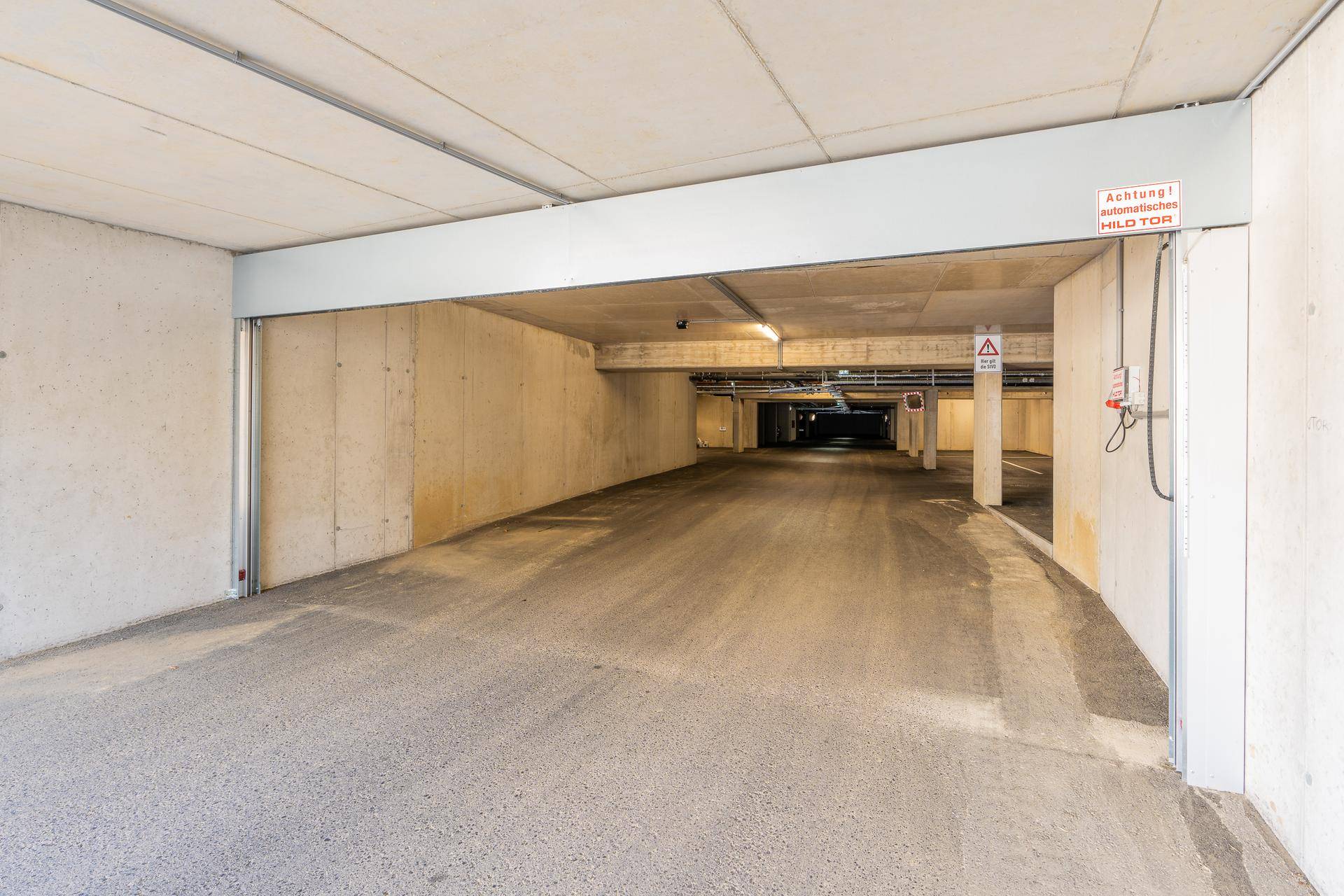 Einfahrt zur Tiefgarage mit Betonwänden, automatischer Schranke und sichtbaren Installationen an der Decke.