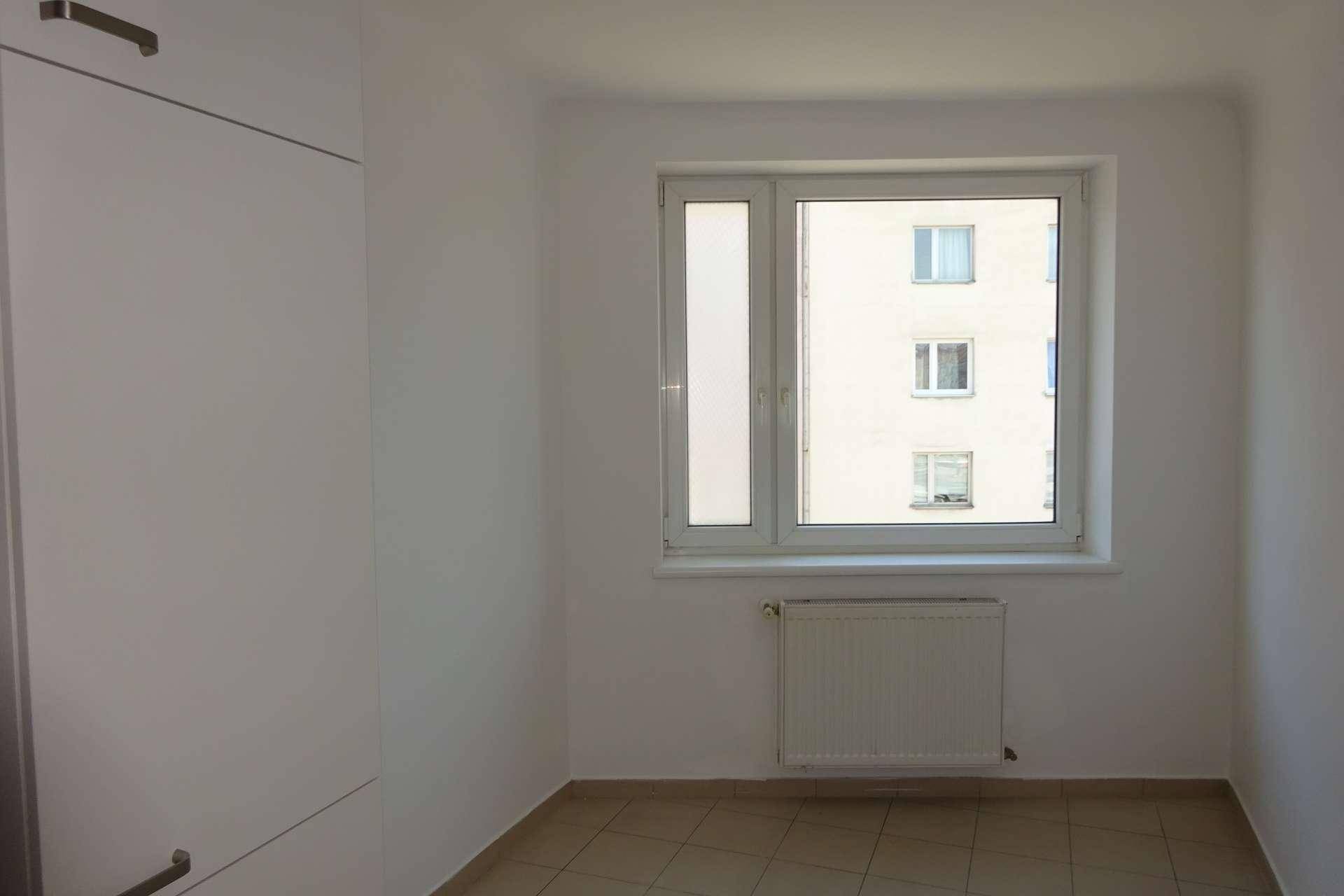 Kleineres Zimmer mit Fliesenboden und einem Fenster, das auf ein gegenüberliegendes Gebäude blickt.