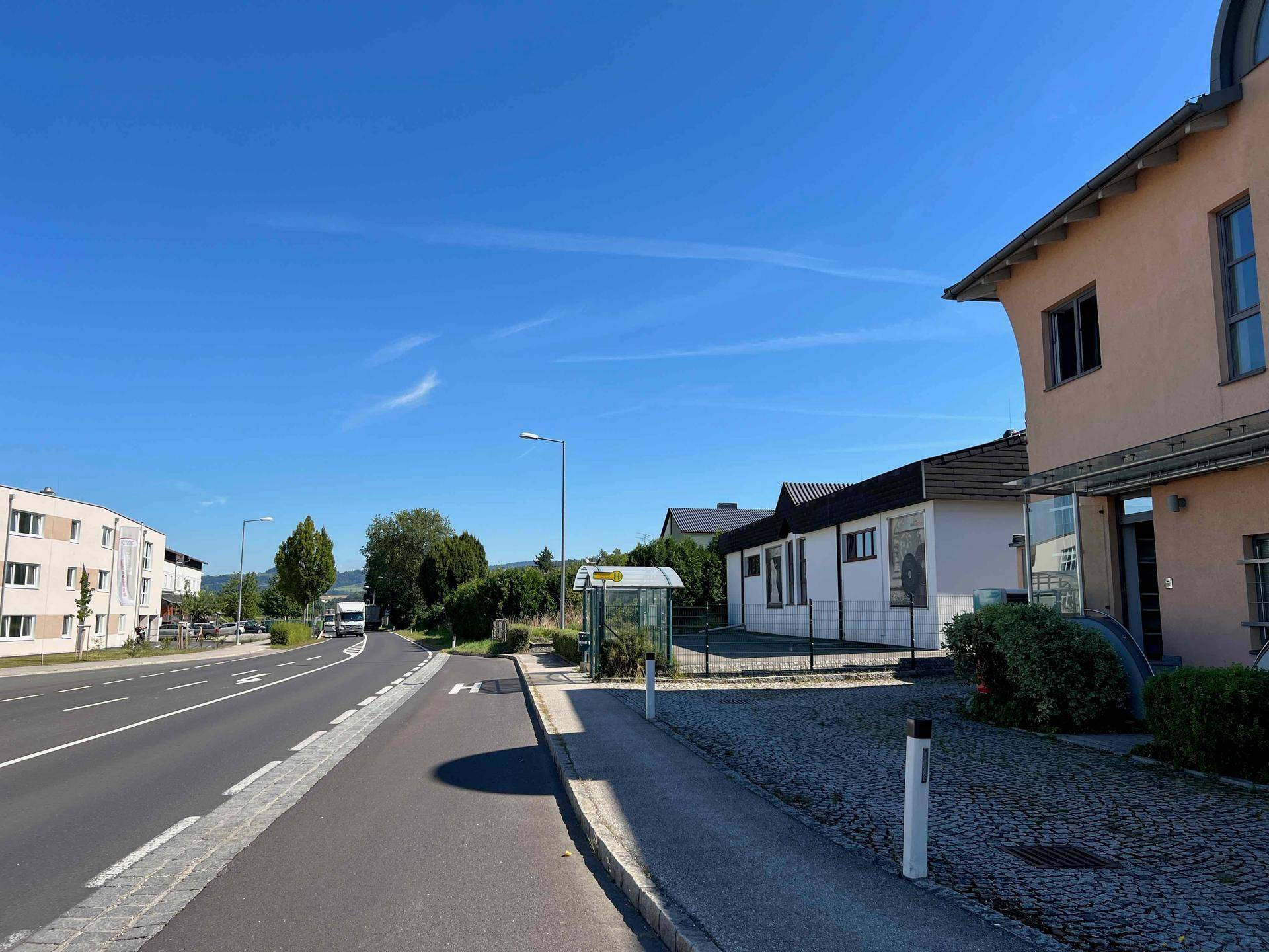 Blick auf eine Straße mit angrenzenden Gebäuden, einem Buswartehäuschen und Bäumen im Hintergrund unter blauem Himmel.