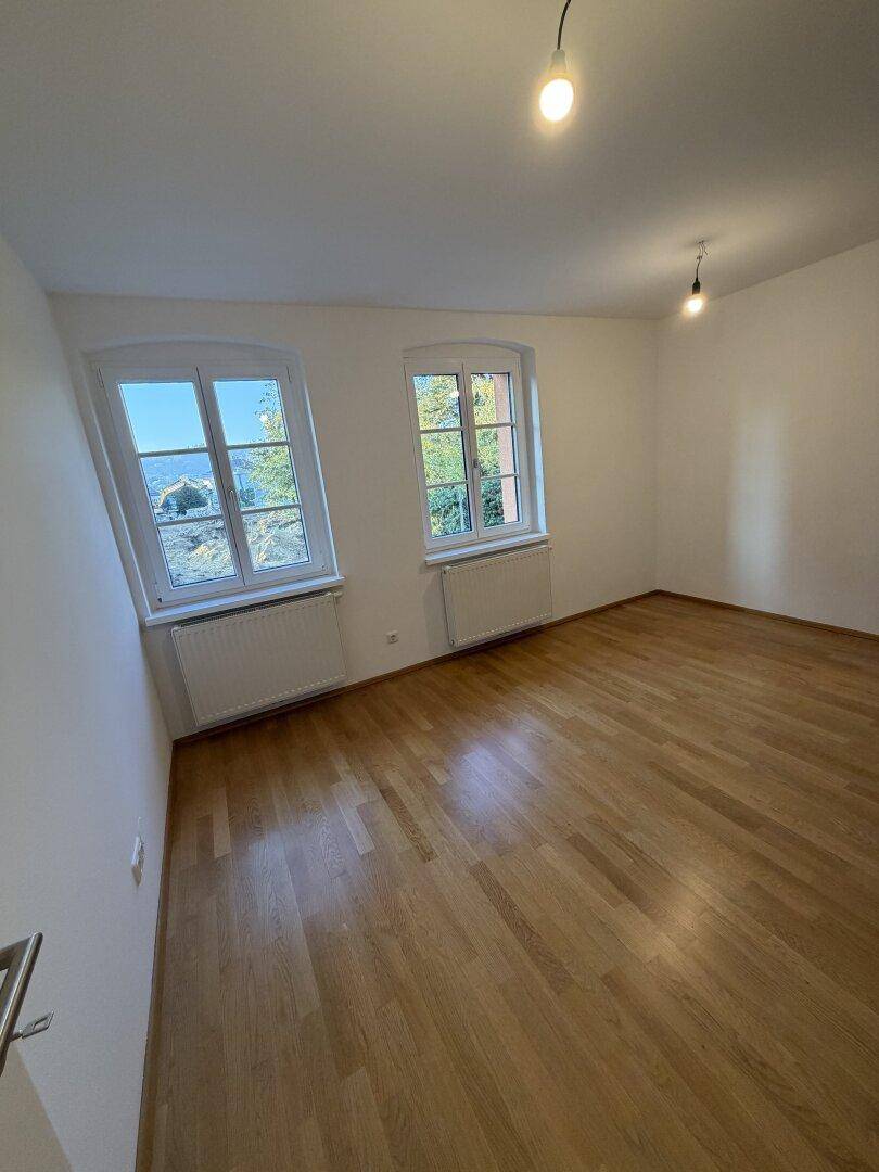 Helles Zimmer mit Holzfußboden und zwei Fenstern, die viel Tageslicht hereinlassen.