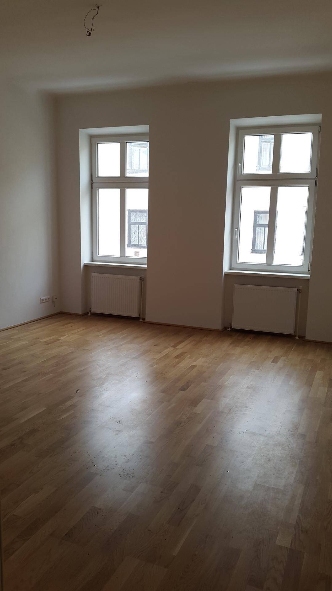 Geräumiges Zimmer mit Holzboden, hohen Decken und zwei großen Fenstern für viel Tageslicht.