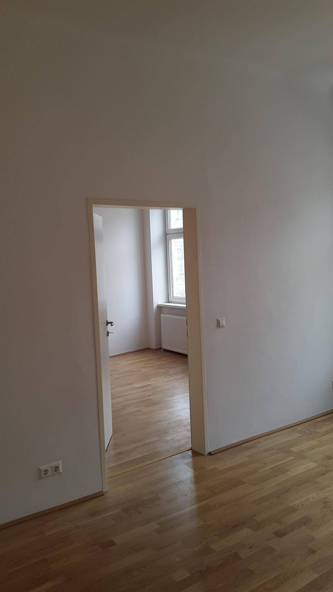 Helles Zimmer mit Holzboden und einem offenen Durchgang zu einem weiteren, lichtdurchfluteten Raum.