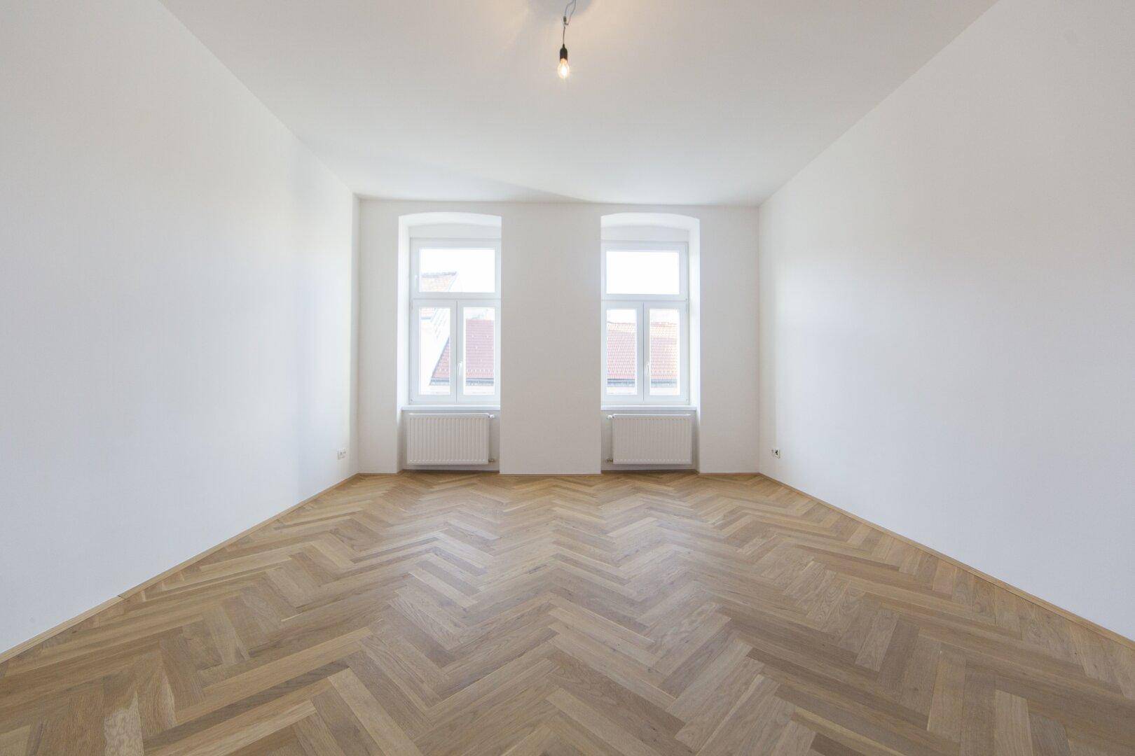 Helles Zimmer mit Fischgrätparkettboden und zwei Fenstern, bereit zur individuellen Gestaltung.