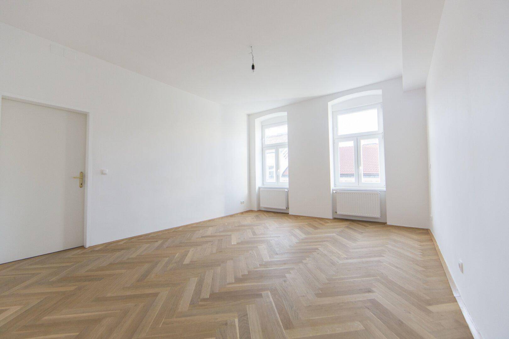 Geräumiges Zimmer mit Fischgrätparkett und weiß gestrichenen Wänden, ideal für Wohnzwecke.