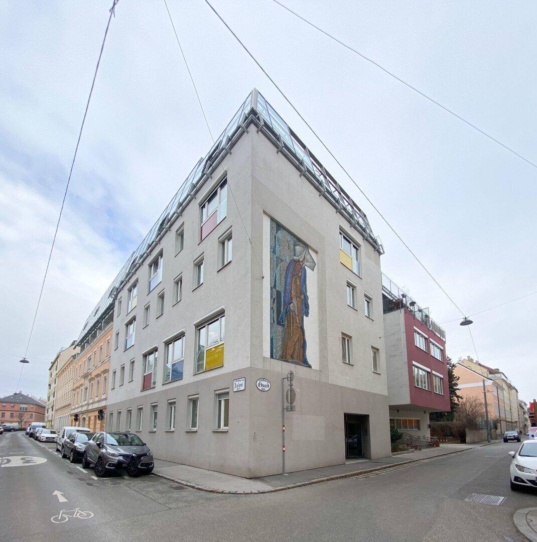 Modernes Eckgebäude mit Glasdach und farbigen Akzenten, integriert in eine städtische Umgebung mit klaren Linien.