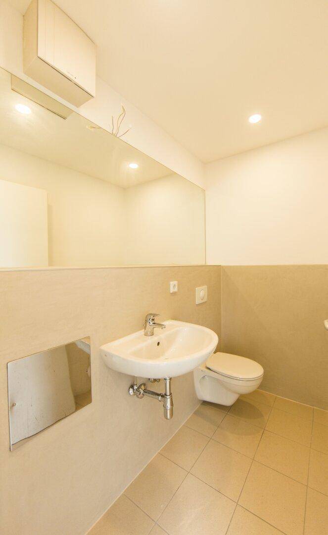 Modernes Badezimmer mit Wand-WC, Waschbecken und großem Spiegel, in hellen Beigetönen gehalten, funktional und sauber.