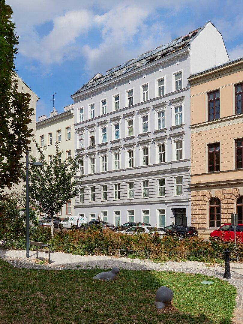 Historisches Gebäude mit heller Fassade und einem kleinen Park im Vordergrund.