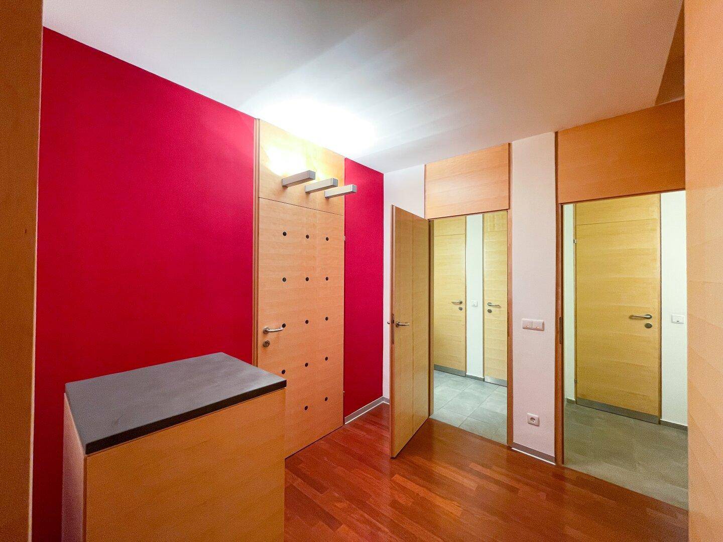 Raum mit auffälliger roter Wand, Holzelementen und mehreren Türen, die zu angrenzenden Bereichen führen.