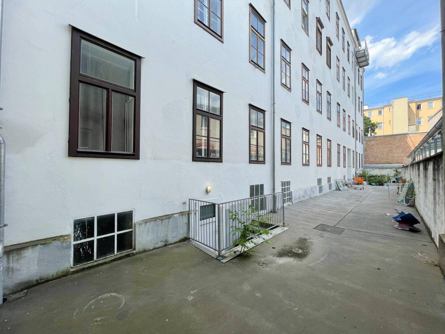 Hinterhof eines Gebäudes mit weißer Fassade, braunen Fenstern und einem Zugang zum Keller.