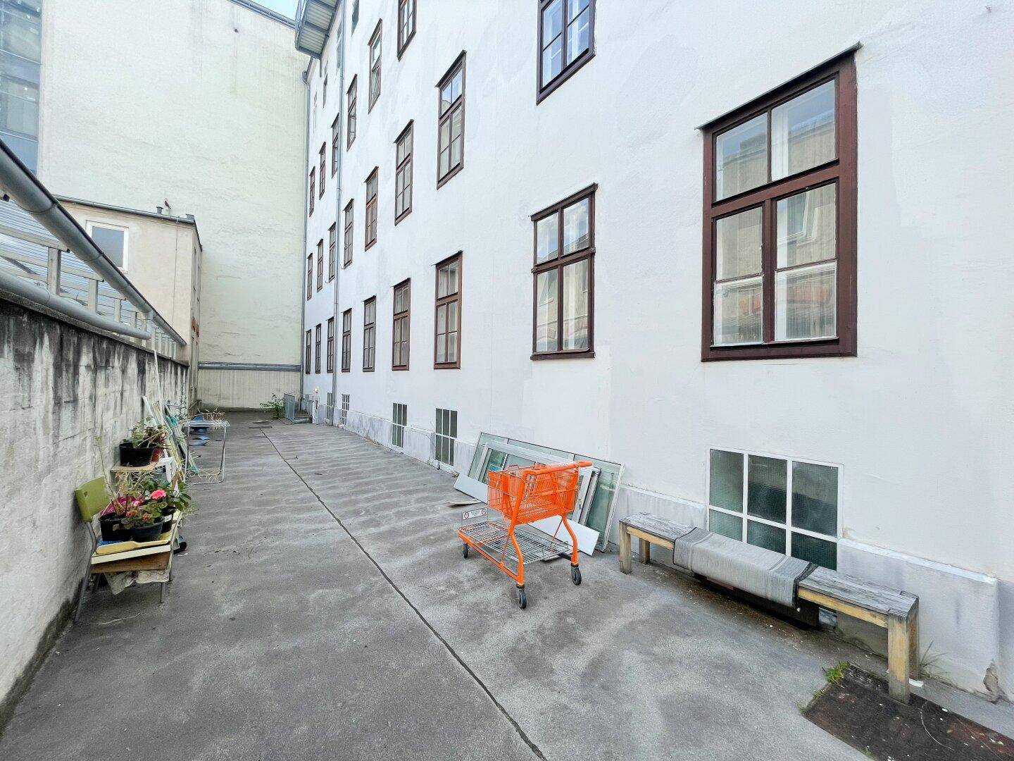 Hinterhof eines Gebäudes mit weißer Fassade, braunen Fenstern und einem schmalen Durchgang.