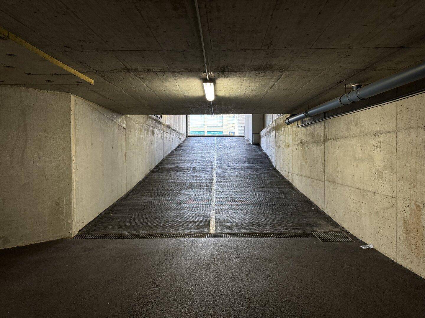 Steile Tiefgaragenrampe mit Betonwänden und Deckenbeleuchtung, die zum Ausgang führt.