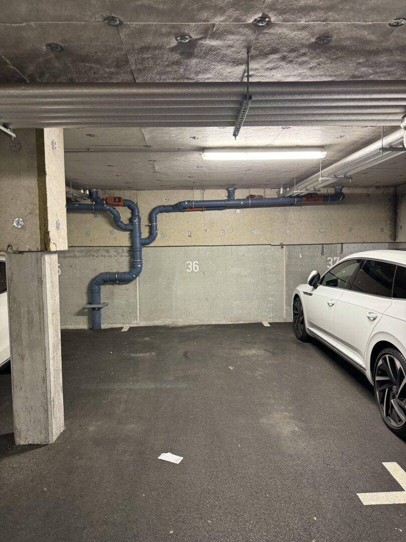 Tiefgarage mit Betonwänden und -decken, gekennzeichneten Stellplätzen und sichtbaren Versorgungsleitungen.