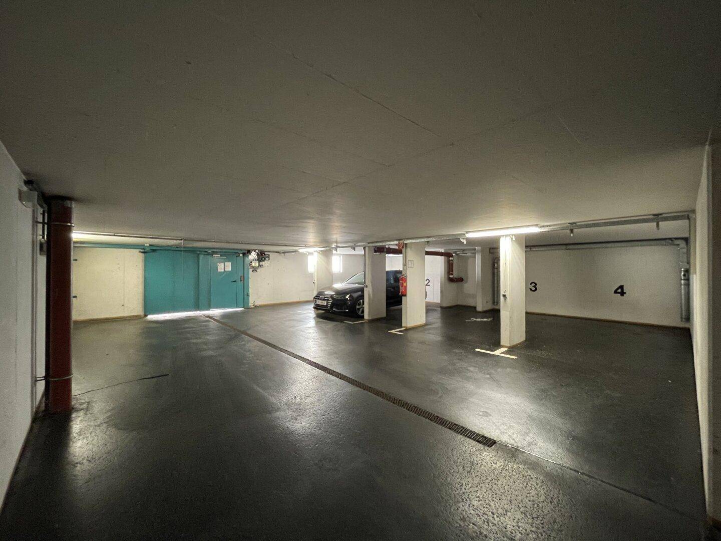 Weitläufige Tiefgarage mit markierten Stellplätzen, einem geparkten Auto und der markanten türkisfarbenen Tür.