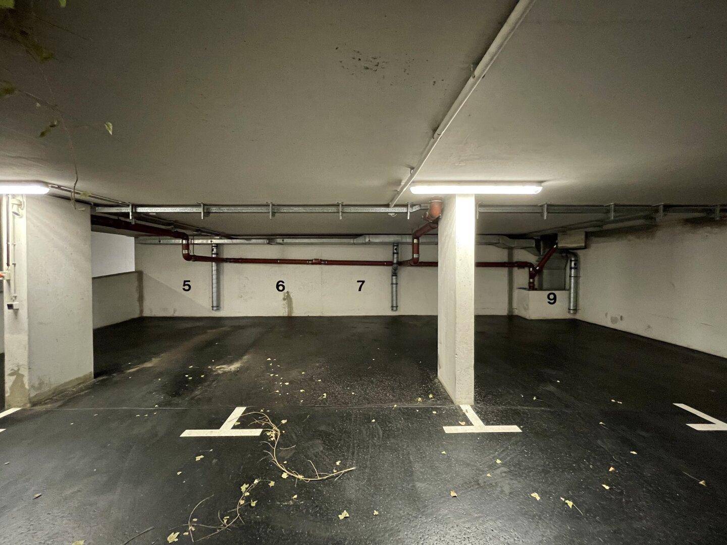 Tiefgarage mit nummerierten Stellplätzen, sichtbaren Rohren an der Decke und einigen Blättern auf dem Boden.