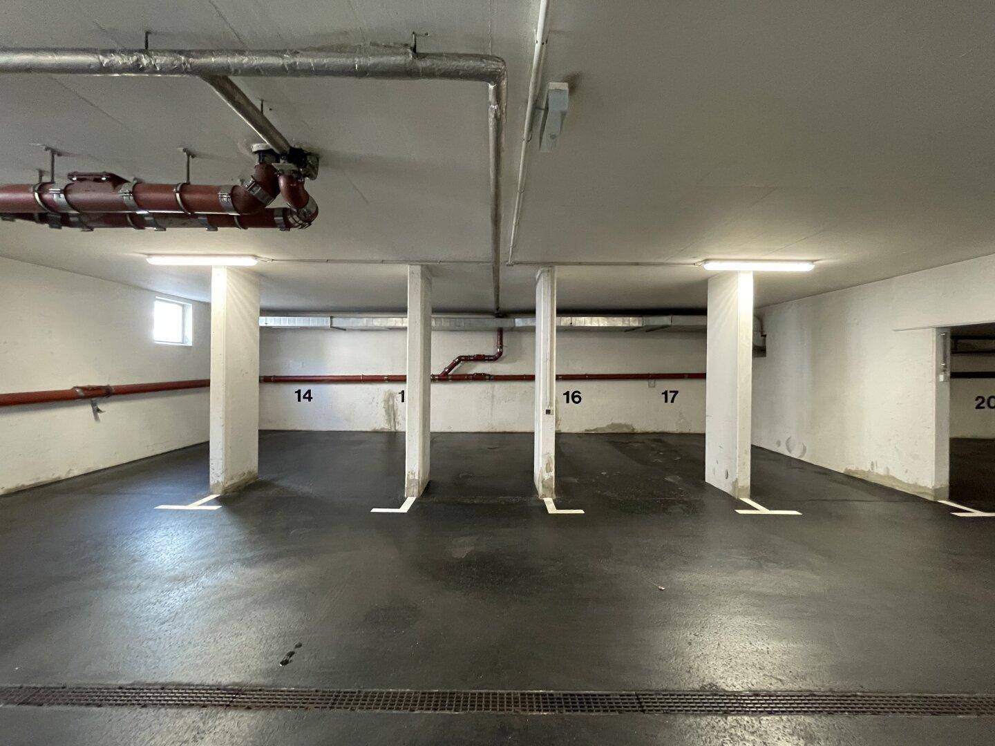 Mehrere nummerierte Stellplätze in einer Tiefgarage mit kleinen Fenstern und sichtbaren Rohren an der Decke.