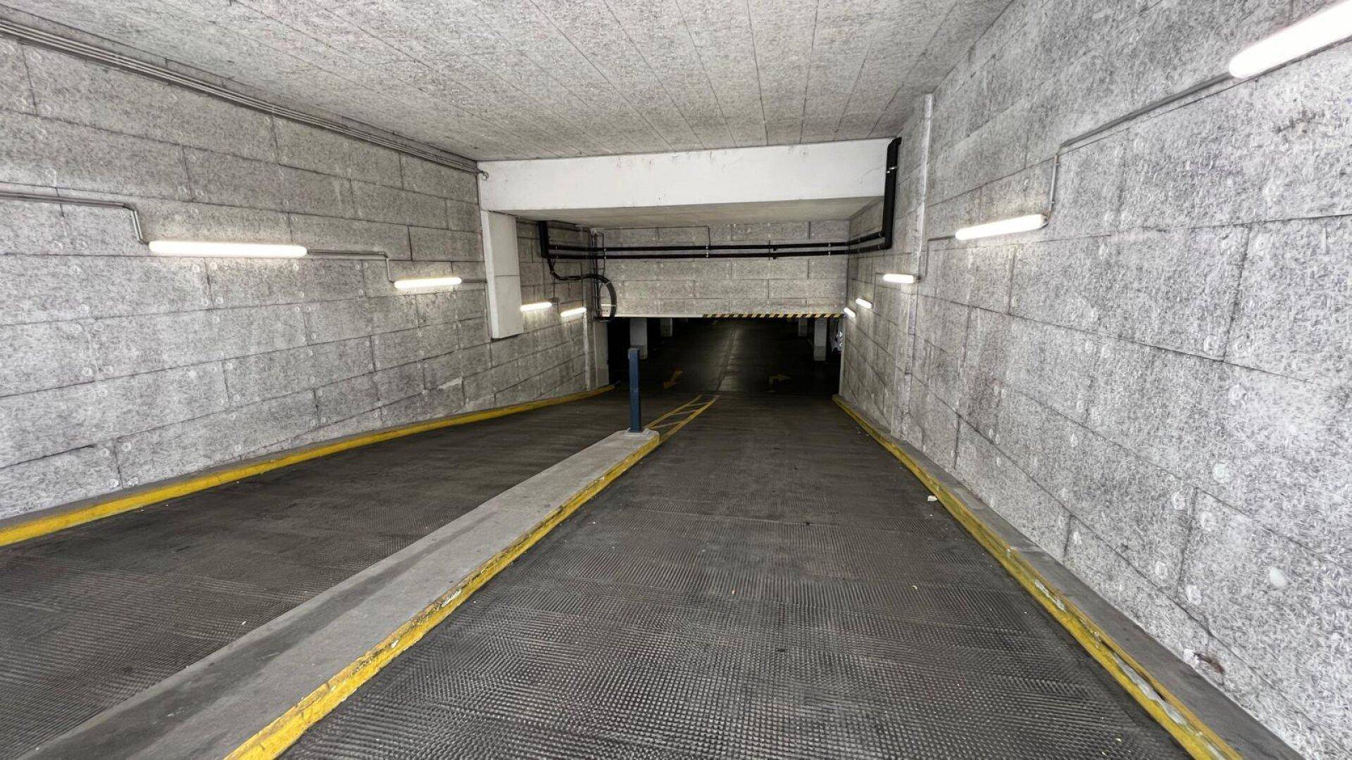 Einfahrt zu einer Tiefgarage mit einer breiten Rampe, grauen Betonwänden und Deckenbeleuchtung.