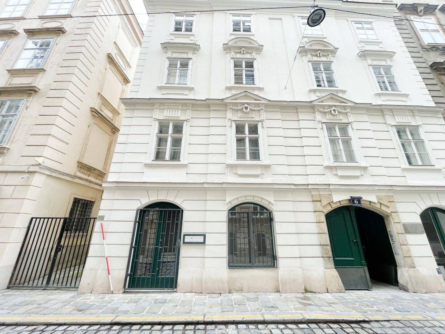 Historische weiße Fassade eines Gebäudes mit mehreren Fenstern und zwei markanten grünen Eingangstüren auf Straßenniveau.