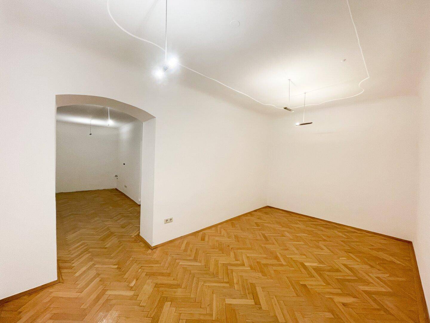 Heller Wohnraum mit Fischgrätparkett und einem eleganten Rundbogen, der zu einem weiteren Zimmer führt.