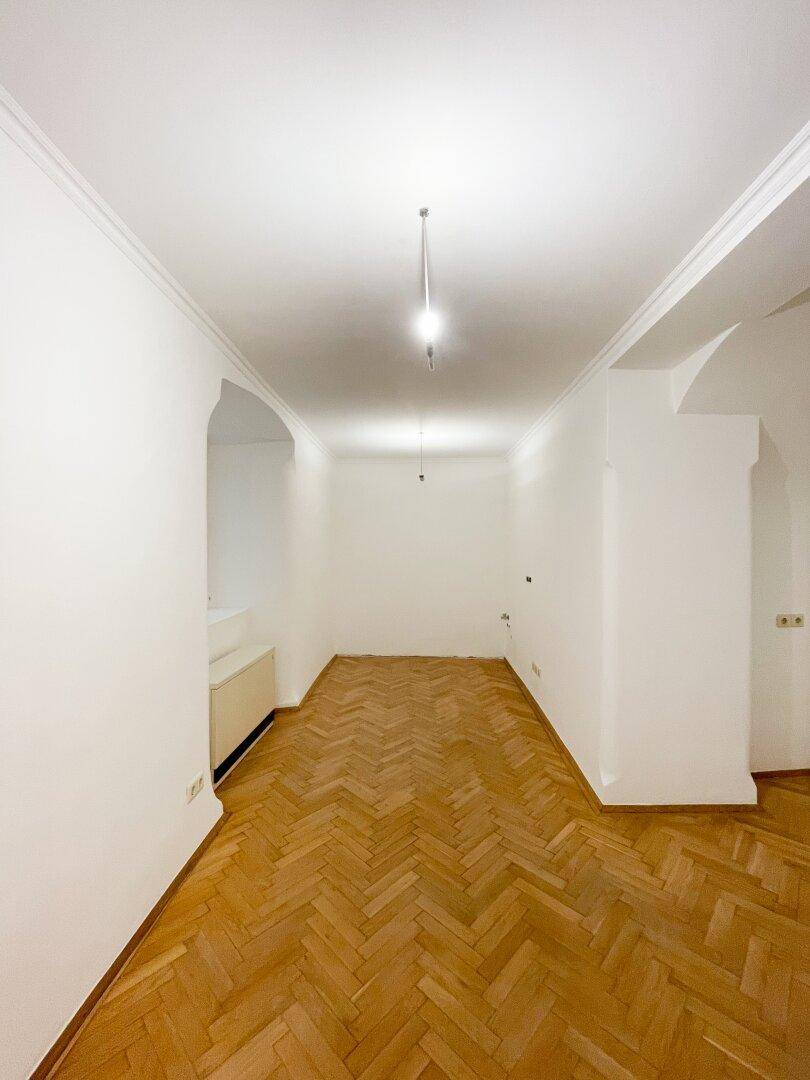 Langer, heller Raum mit Fischgrätparkett und schlichten weißen Wänden, ideal für vielseitige Nutzung.