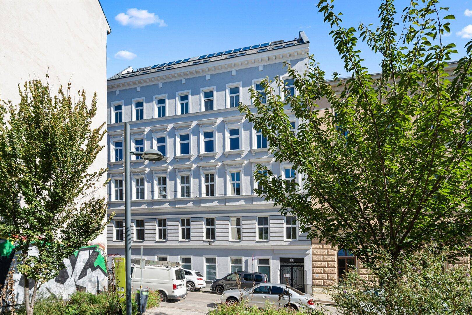 Historisches Gebäude mit blauer Fassade, vielen Fenstern und Bäumen im Vordergrund.