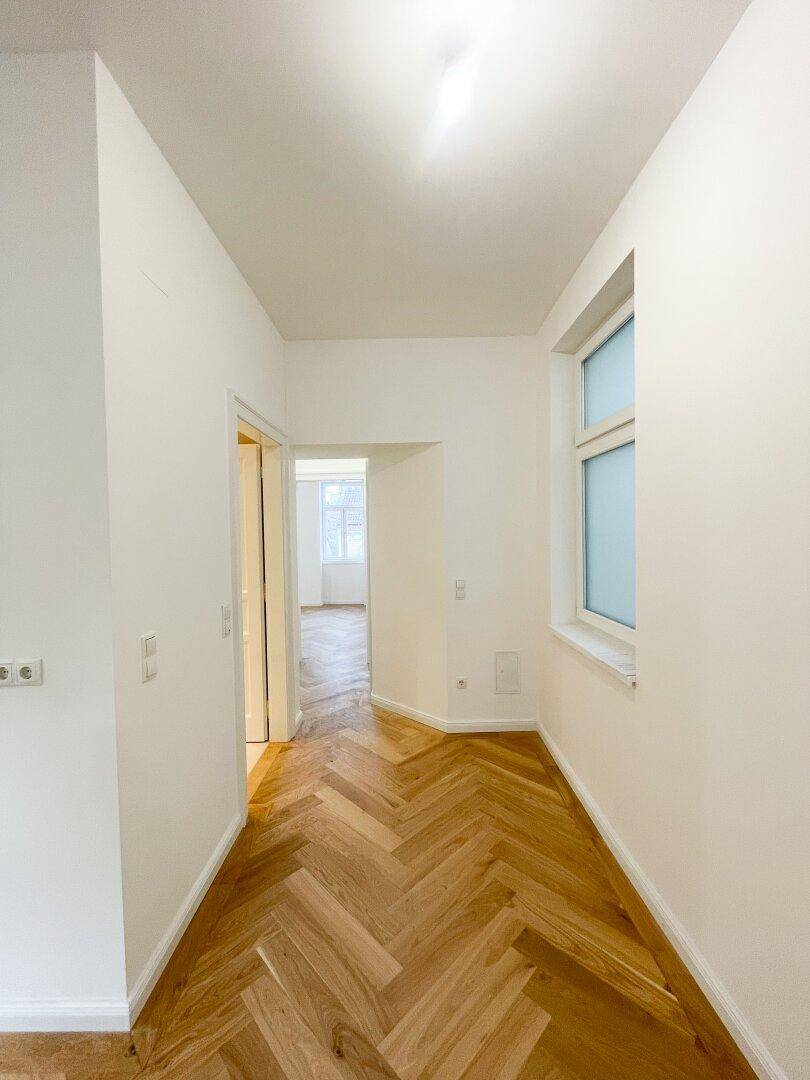 Langer, heller Flur mit Fischgrätparkett und einem Fenster am Ende, das Tageslicht spendet.