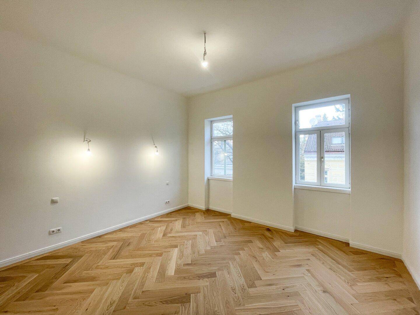 Helles Zimmer mit zwei Fenstern und elegantem Fischgrätparkett, bereit zur individuellen Gestaltung.
