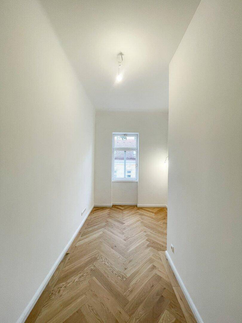 Schmaler Flur mit Fischgrätparkett und einem Fenster am Ende, das den Raum erhellt.