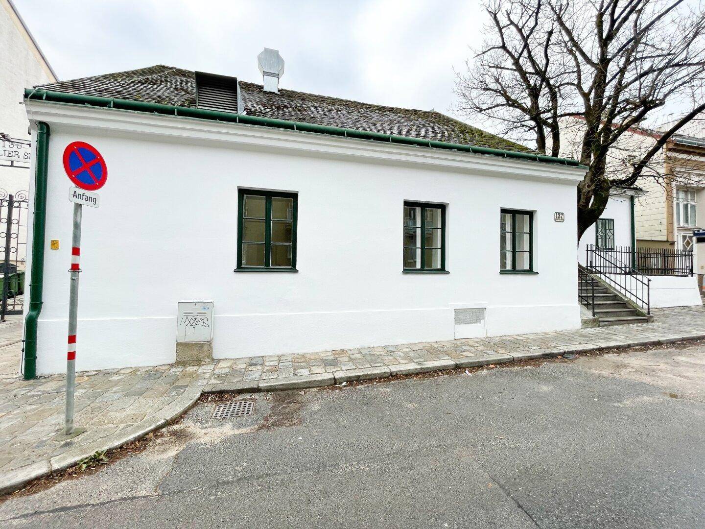 Weiß verputztes Gebäude mit grünen Fensterrahmen und einem traditionellen Dach, an einer ruhigen Straße gelegen.