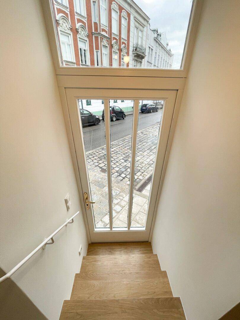 Heller Treppenaufgang mit Holzstufen und einem Fenster, das den Blick auf die Kopfsteinpflasterstraße freigibt.
