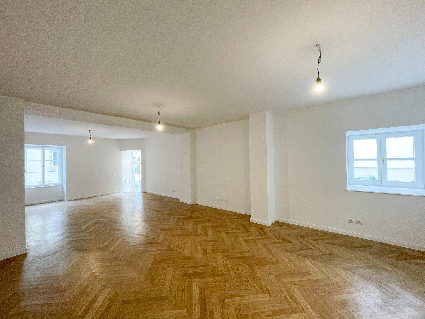 Heller Wohnraum mit klassischem Fischgrätparkett und mehreren Fenstern, die viel Tageslicht hereinlassen.