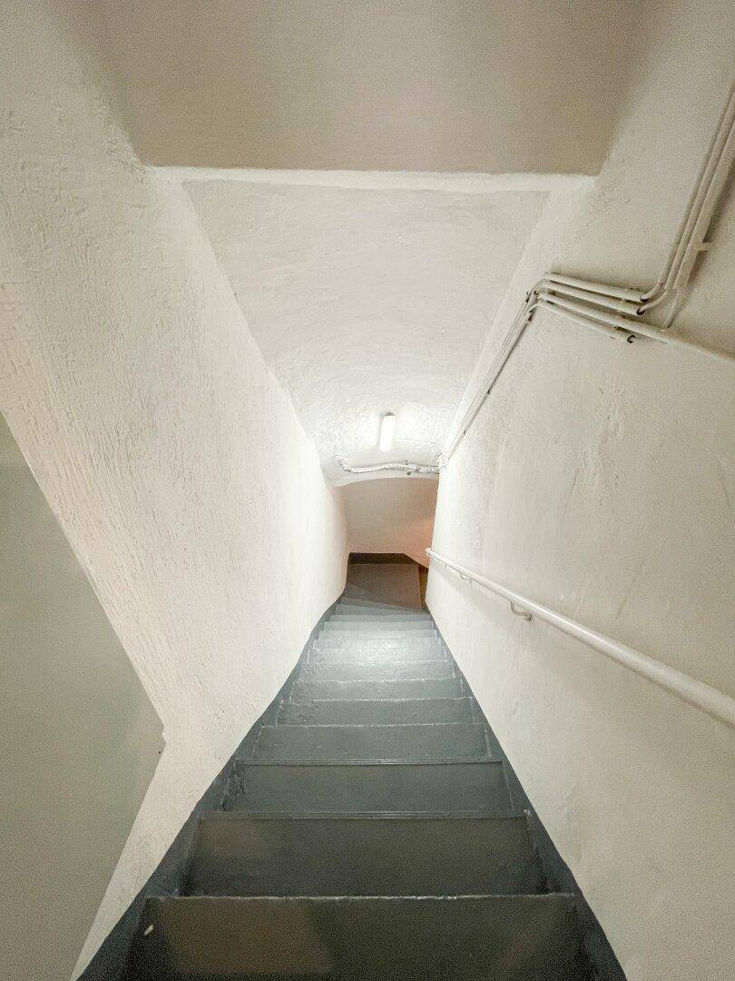 Steile Kellertreppe mit grau gestrichenen Stufen und einem einfachen Handlauf, der in den Keller führt.